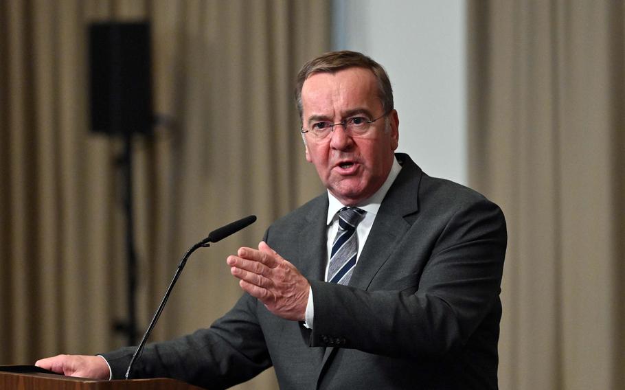 German Defense Minister Boris Pistorius holds out his hand as he speaks into a microphone at a lecturn.