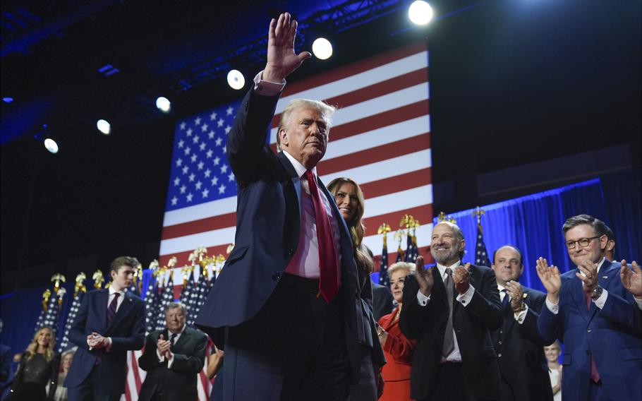 Donald Trump waves to the crowd while standing on stage in front of supporters and large American flag.