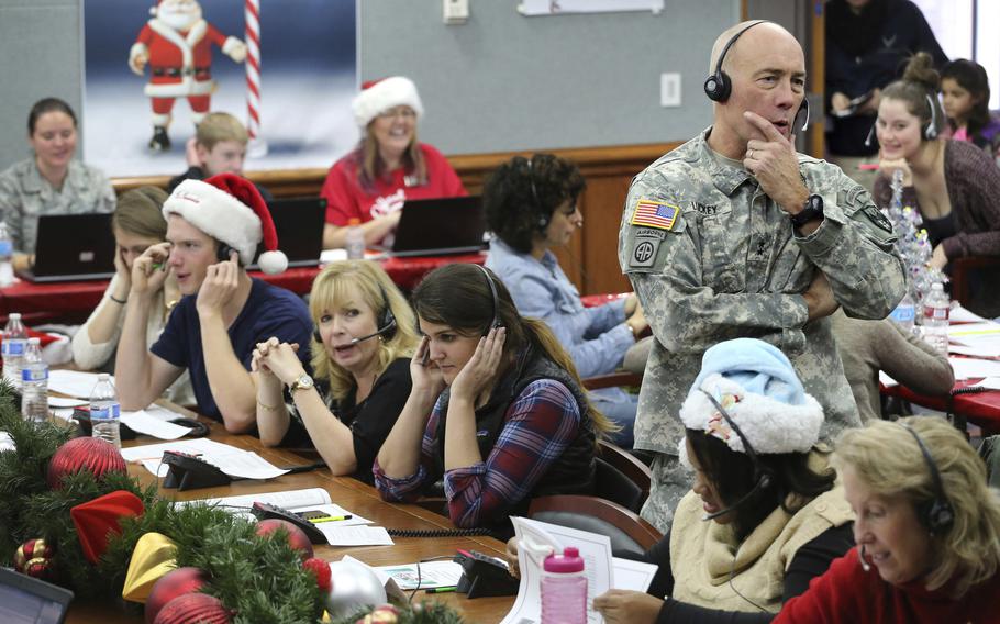 A soldier in camouflage uniform wearing a radion headset stands next to a long table of volunteers wearing Santa hats and radio headsets.