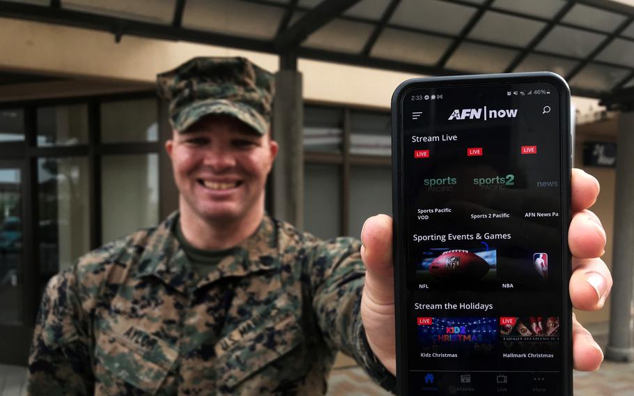 A Marine in uniform smiles as he holds out his phone with the AFN app on the screen.