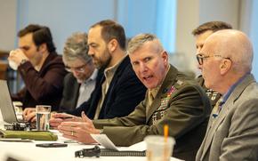 A Marine Corps general in uniform speaks and sits in the middle of a long table with other men seated on either side.