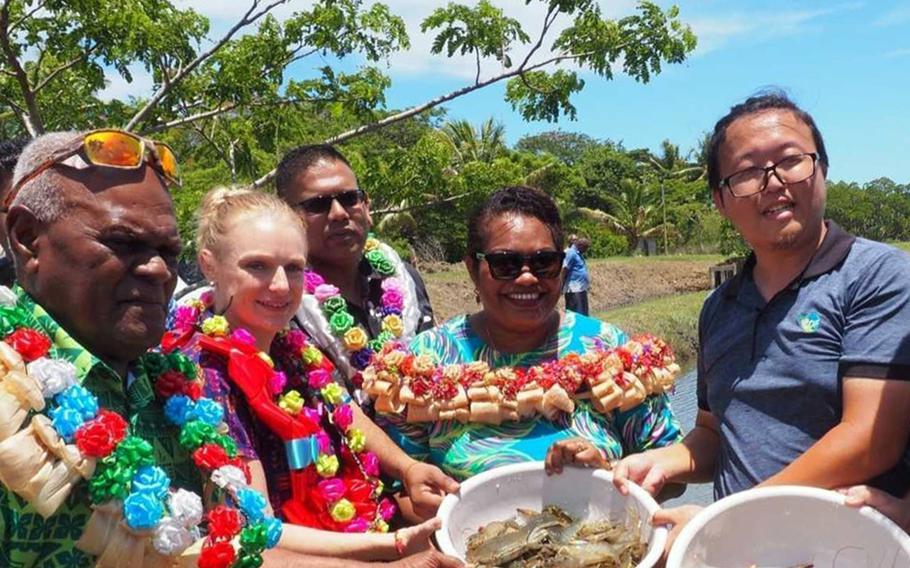 People in floral shirts and flower leis hold a bowl of shrimp on a beach with palm trees in the background.