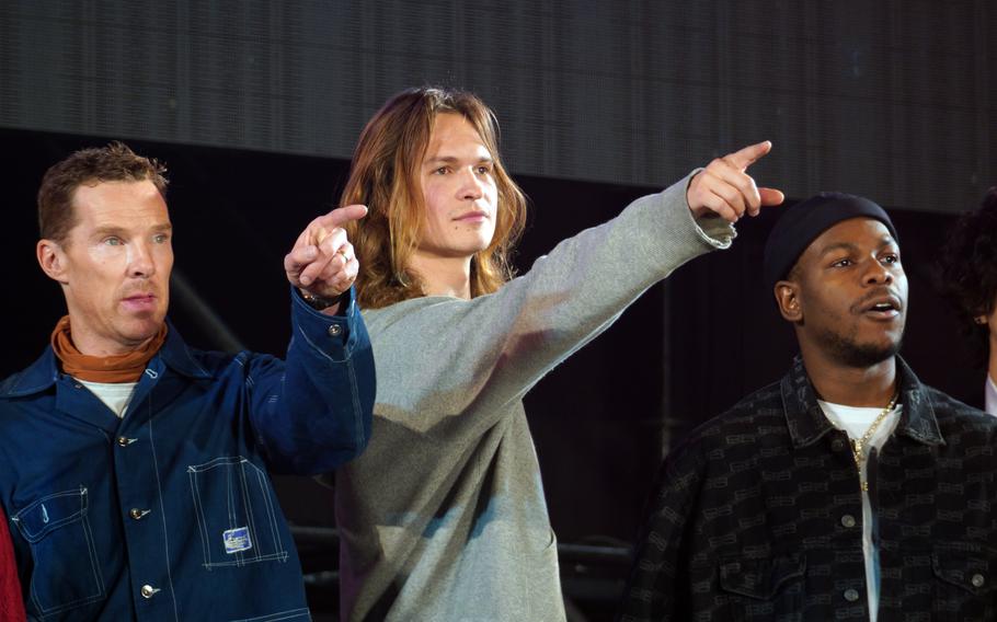 Three men stand on a stage, with two pointing out towards the audience.