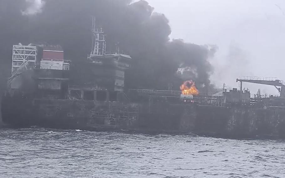 Black smoke billows from a cargo ship as it sits in open water and a small fire blazes on the deck.