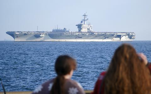 An aircraft carrier is in the background and two women, with their backs to the camera, are in the foreground looking in the direction of the ship from a pier.