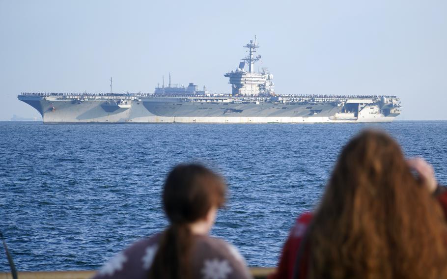 An aircraft carrier is in the background and two women, with their backs to the camera, are in the foreground looking in the direction of the ship from a pier.