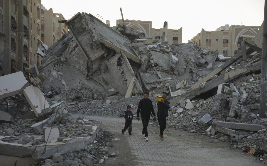 Two men and a boy walk along a path past heaped rubble of destroyed buildings.