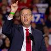 A man in a suit and glasses gestures to the crowd while standing at a podium and speaking into a microphone at a campaign event.