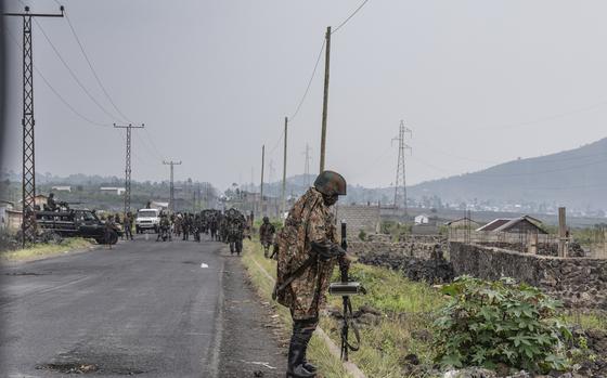 A soldier stands at the side of a road and adjusts his gun, while a larger group of soldiers is seen in the background.