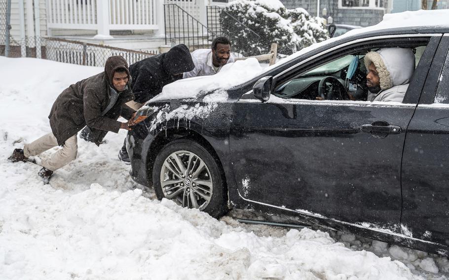 A man steps on the gas of his car while three other push his car that is stuck in snow.