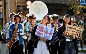 Japanese college students hold signs and play instruments as they walk down a street.