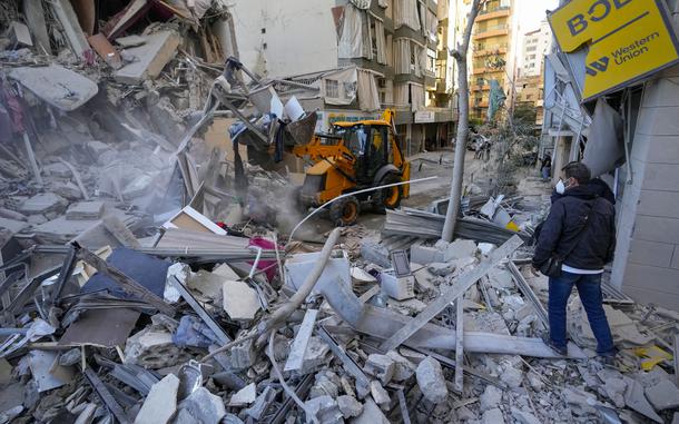 A man in a bulldozer clears rubble from destroyed buildings while others look on.