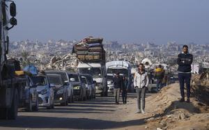 Palestinians stand next to a line of stationary cars on a road with a skyline of destroyed buildings in the background.