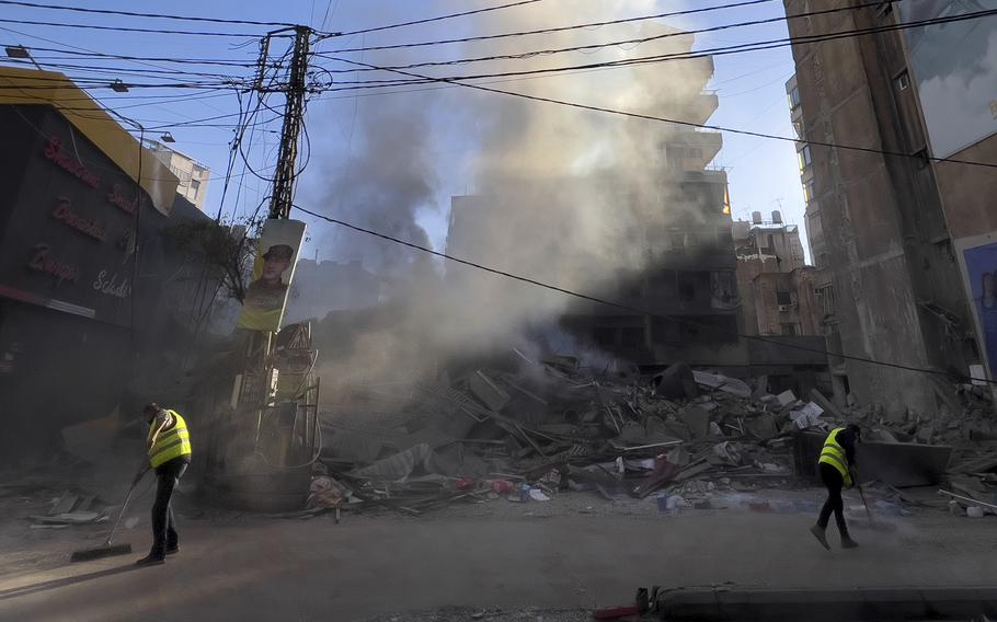 Workers clean a street as smoke rises from a destroyed building.