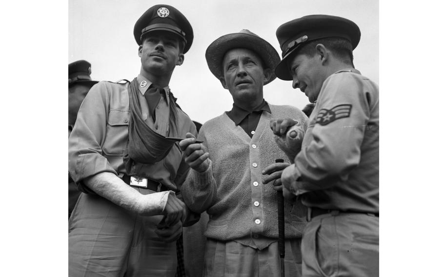 A black-and-white image of Bing Crosby in a button-down sweater standing between two military service members in dress uniforms.