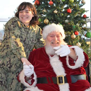 A Navy hospitalman, Rayven Palmer, smiles and takes the hand of a smiling Santa in front of the Christmas tree.