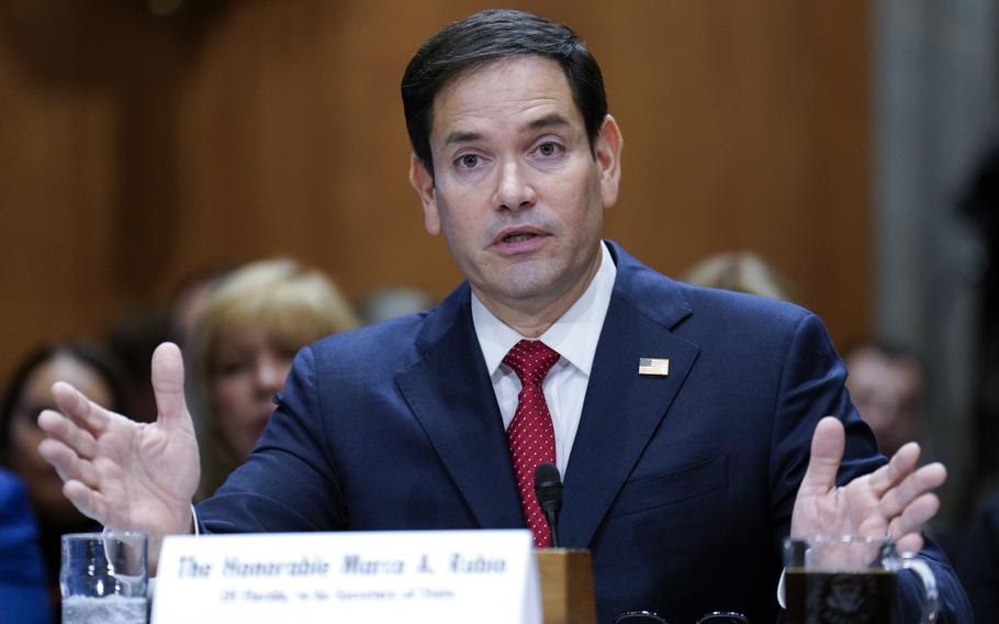 Marco Rubio speaks while seated with his palms stretched outwards; an out-of-focus placard rests on the table in front of him.