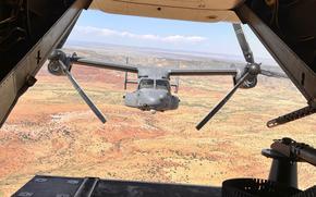 An Osprey military aircraft is seen from the back of another Osprey as they fly in formation over a desert setting.