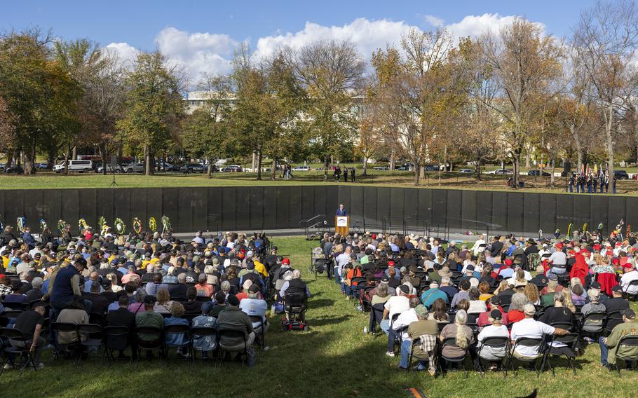 A crowd of people sits in front of a veterans memorial while listening to a speaker.