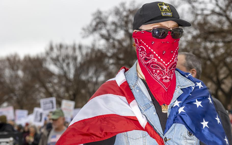 A man in an Army baseball cap, wearing sunglasses and a bandanna over his face with the American flag draped over his shoulders.