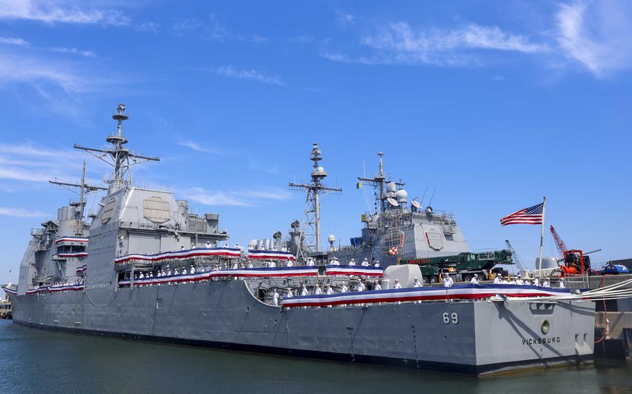 A Navy guided-missile cruiser is lined with sailors and streamers on the rails.