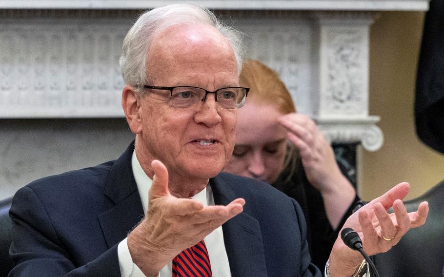 A seated congressman speaks into a microphone during a meeting while raising both hands with his palms up.