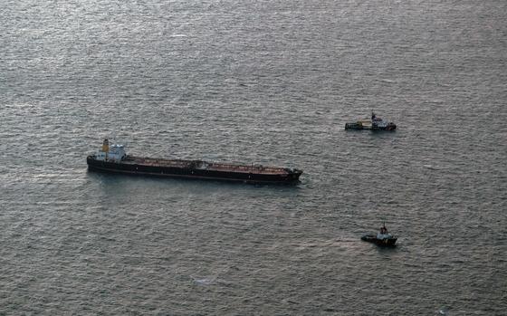 A tanker at sea is seen from above, with two small tugboats in front of it.