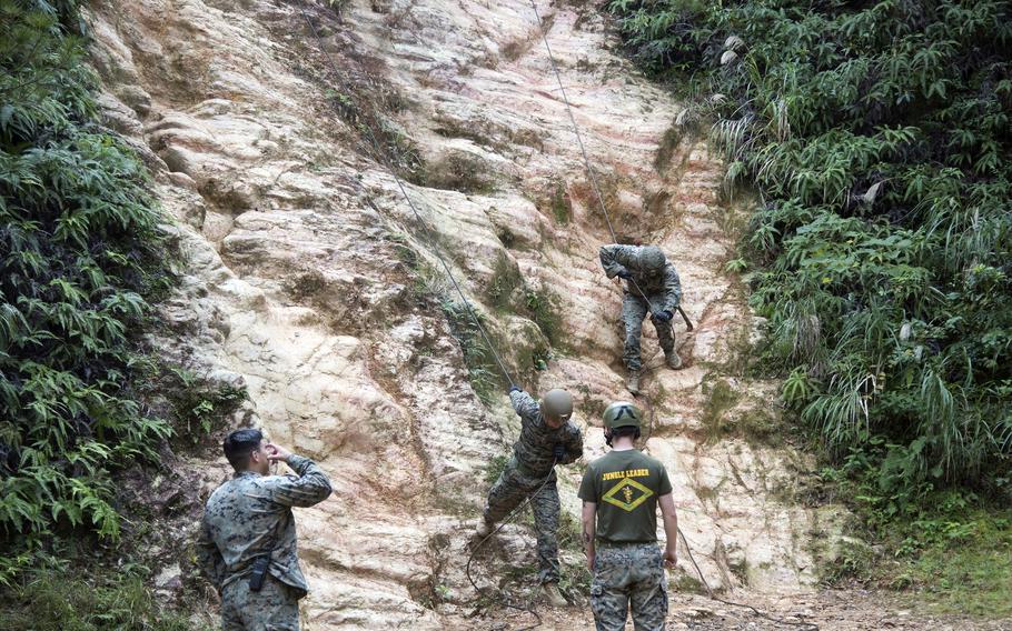 Two Marines stand at the base of a cliff as two others descend it on ropes.