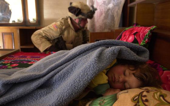 A young child sleeps under a blanket while a soldier in combat gear conducts a search in the background.