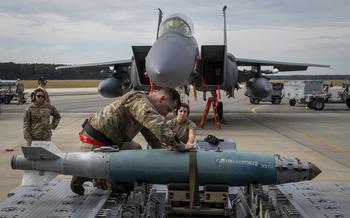 A U.S. airmen kneels on top of an MJ-1 bomb lift truck as he straps down a GBU-38 munition on an airfield in 2021.