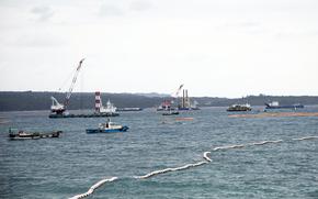 Construction vessels sit in open water with a coastline in the background.