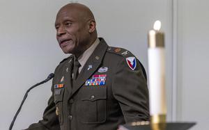 A man in uniform speaking at a podium with a white candle in the foreground to his left.