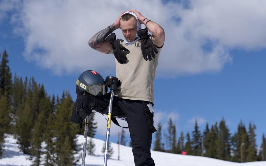 A man with a prosthetic leg pulls a winter covering over his head while standing on a snowy mountin with evergreen trees and cloudy blue sky in the background.