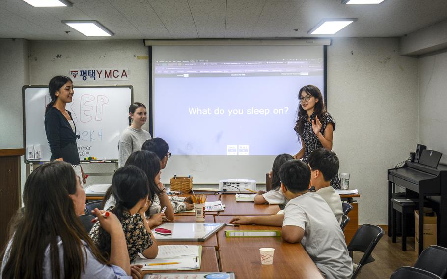 A teenage girl stands in front of a screen with “What do you sleep on?” projected on it as she teaches an English class.