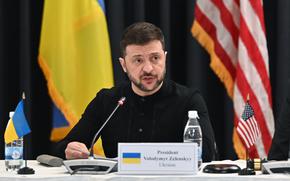 Ukrainian President Volodymyr Zelenskyy sits at a table with a A Ukrainian flag and a U.S. flag behind him. 