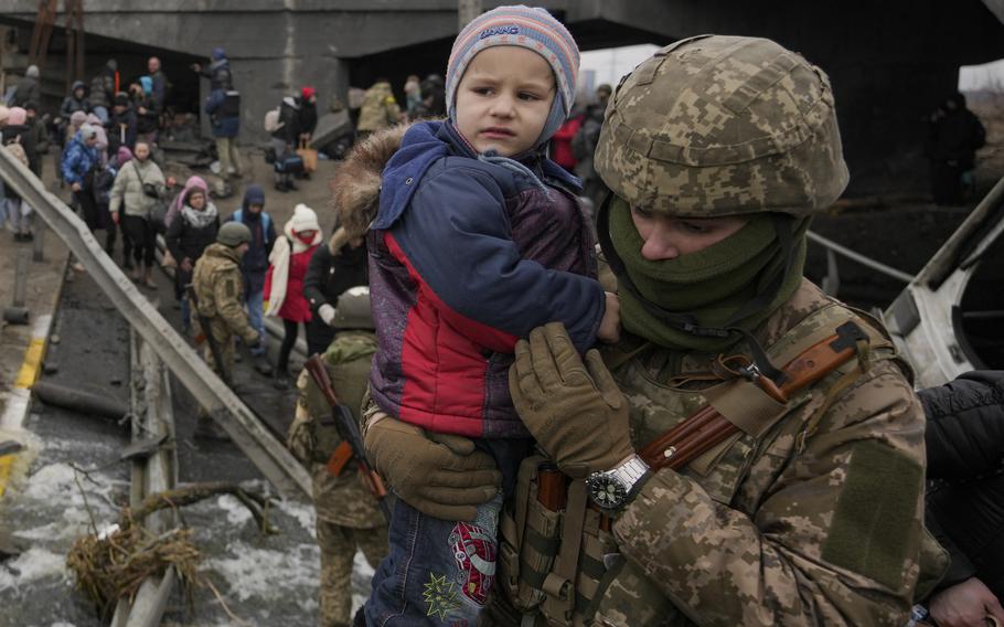 A Ukrainian military service member in uniform carries a child wearing warm clothing.