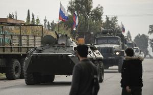 Two people watch as armored vehicles with Russian flags attached roll by.