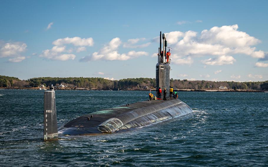 A submarine travels through open water with its top half visible above the water and a coast line in the background.