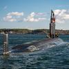 A submarine travels through open water with its top half visible above the water and a coast line in the background.