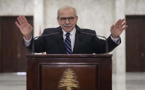 A man in a suit and glasses stands mostly covered by a podium, raising both hands with the palms open.