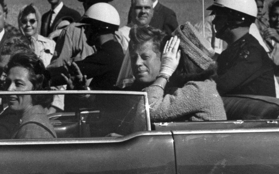 A black-and-white 1963 image shows John F. Kennedy riding in an open-topped car next to his wife Jacqueline Kennedy, who has her hand up obscuring her face.