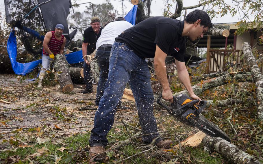 A man uses a chainsaw to cut through branches as he and others clear debris from a yard after a tornado.