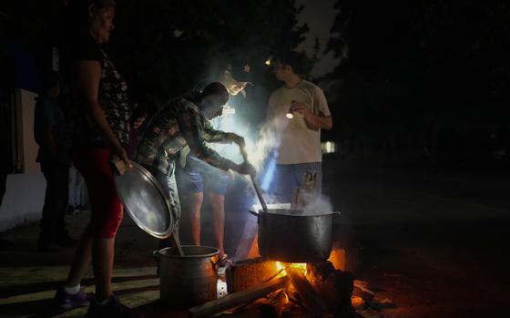 A man outdoors at night stirs a large pot over a fire while another man holds a flashlight on the pot.