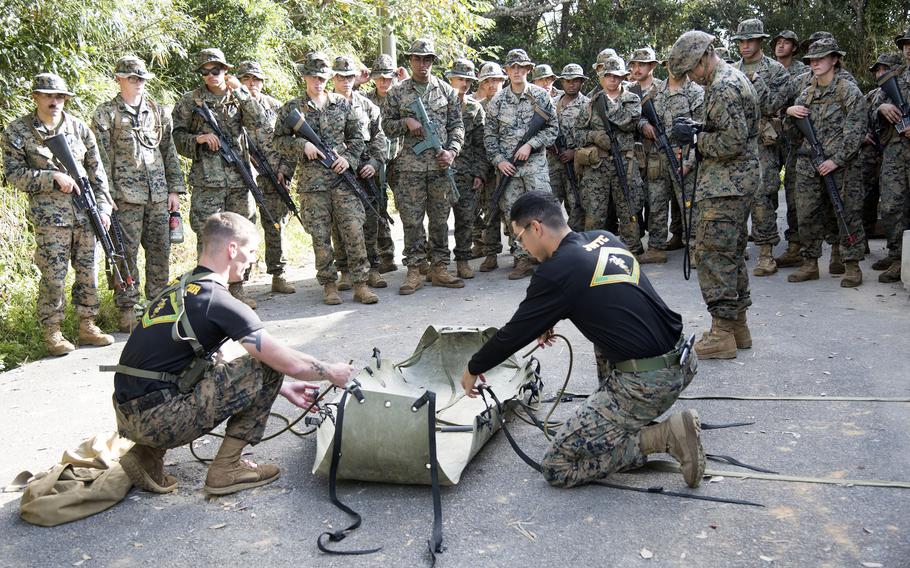 A group of Marines watch as two Marines hold a stretcher. 