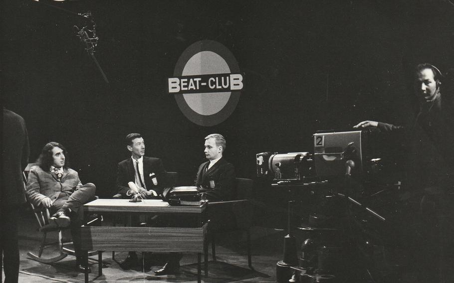 A black and white photo shows three men sat at a table in a television studio.