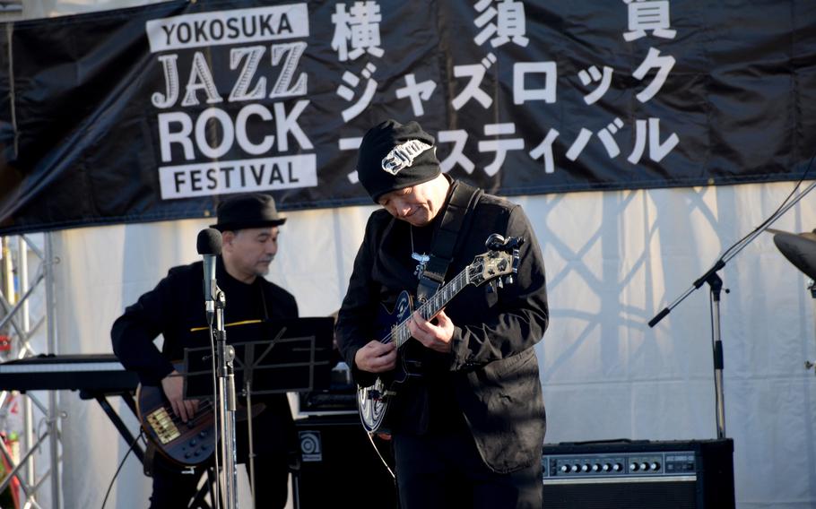 Japanese musicians play guitars on an outdoor stage under a banner for the Yokosuka Jazz Rock Festival.