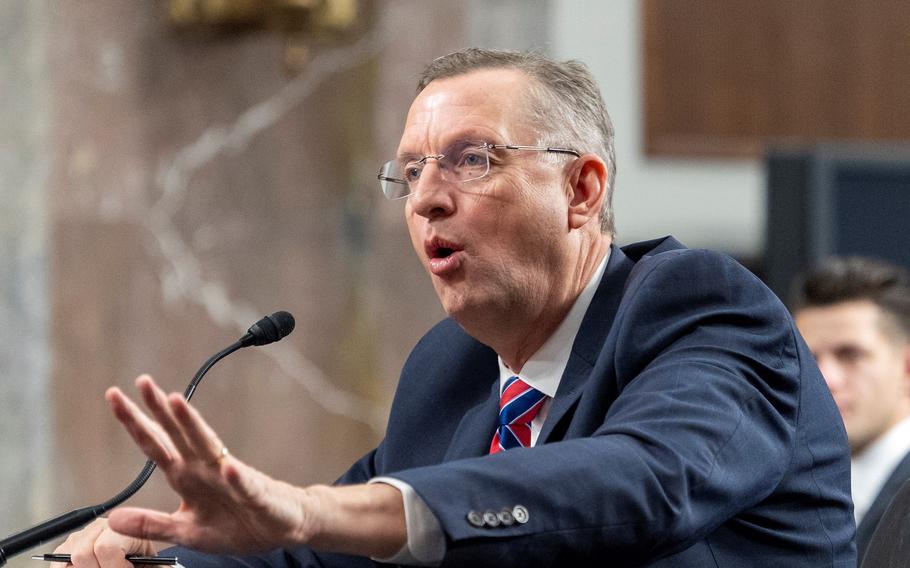 Collins seated, raising his left hand and speaking into a microphone.