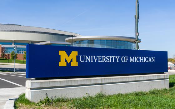 A sign on a patch of grass in front of some buildings that reads “University of Michigan” with the school logo.