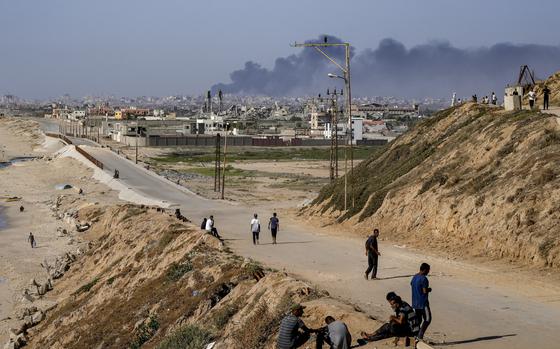 Gazans sit or walk along a road while smoke plumes rise in the background.