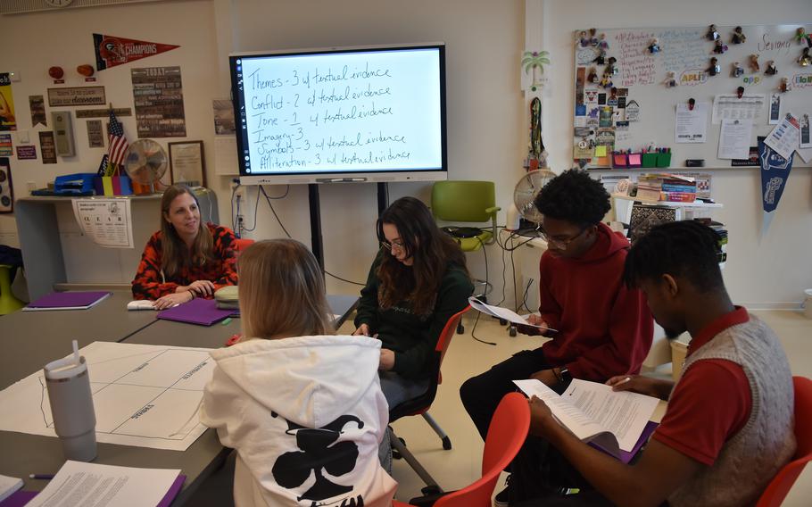 Students and teachers in a classroom, with a video screen in the background.